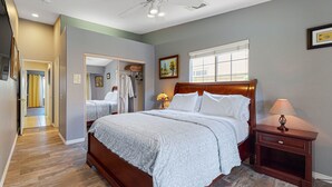 A bedroom with a wooden bed, a nightstand, a window, and a door leading to a bathroom. - Family-Friendly Home w/ an Outdoor Kitchen, Private Pool, & Spa - 3BR - #66708 (La Quinta)
