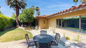 A patio with a round table and chairs, surrounded by palm trees and desert plants. - Siesta-ready home with private saltwater pool - close to downtown (Palm Springs)