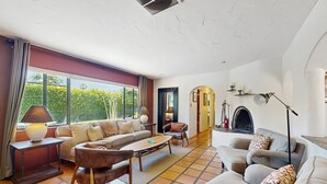 A spacious living room with a large sofa, a coffee table, and a fireplace. There is a window with curtains and a lamp on a side table. - Siesta-ready home with private saltwater pool - close to downtown (Palm Springs)