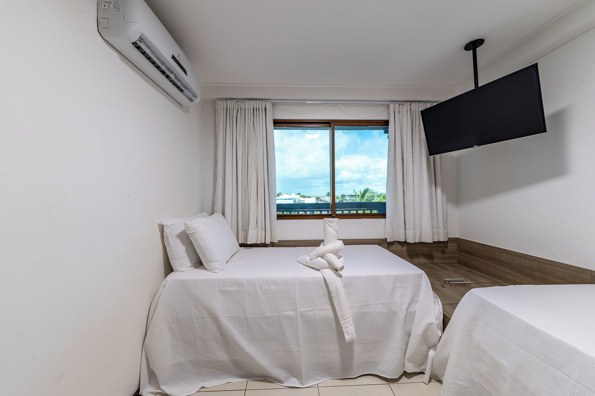 A hotel room with two beds, a flat-screen TV, an air conditioner, and a window with curtains.
