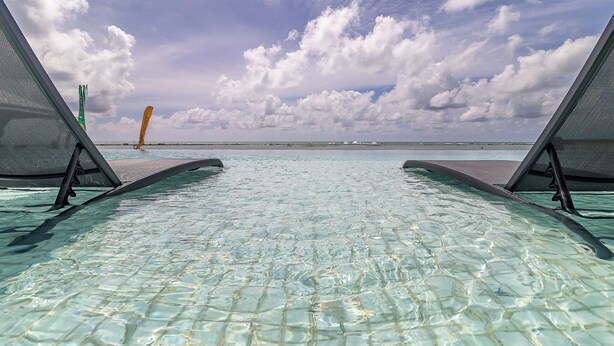 Ein klares Schwimmbecken mit zwei Liegestühlen, die zum Wasser blicken, ein Strand in der Ferne und ein teilweise bewölkter Himmel.