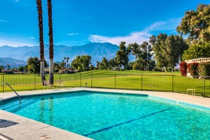 A swimming pool with clear blue water, surrounded by a fenced area with green grass and trees. - home w/ a patio & shared pool plus fairway & mountain views (Rancho Mirage)
