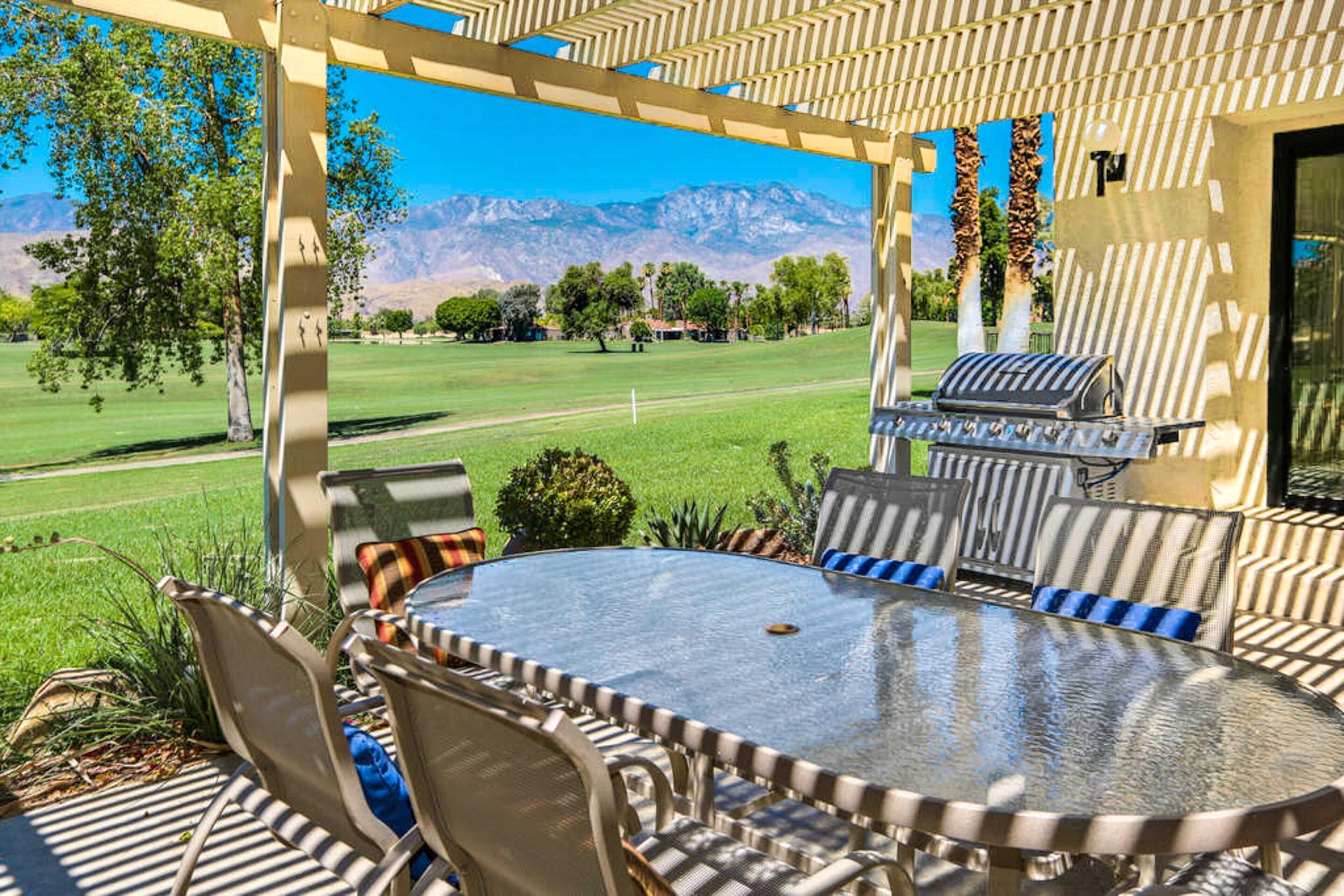 Eine Terrasse im Freien mit einem runden Glastisch, Metallstühlen, einem Grill und Blick auf einen Golfplatz und Berge.