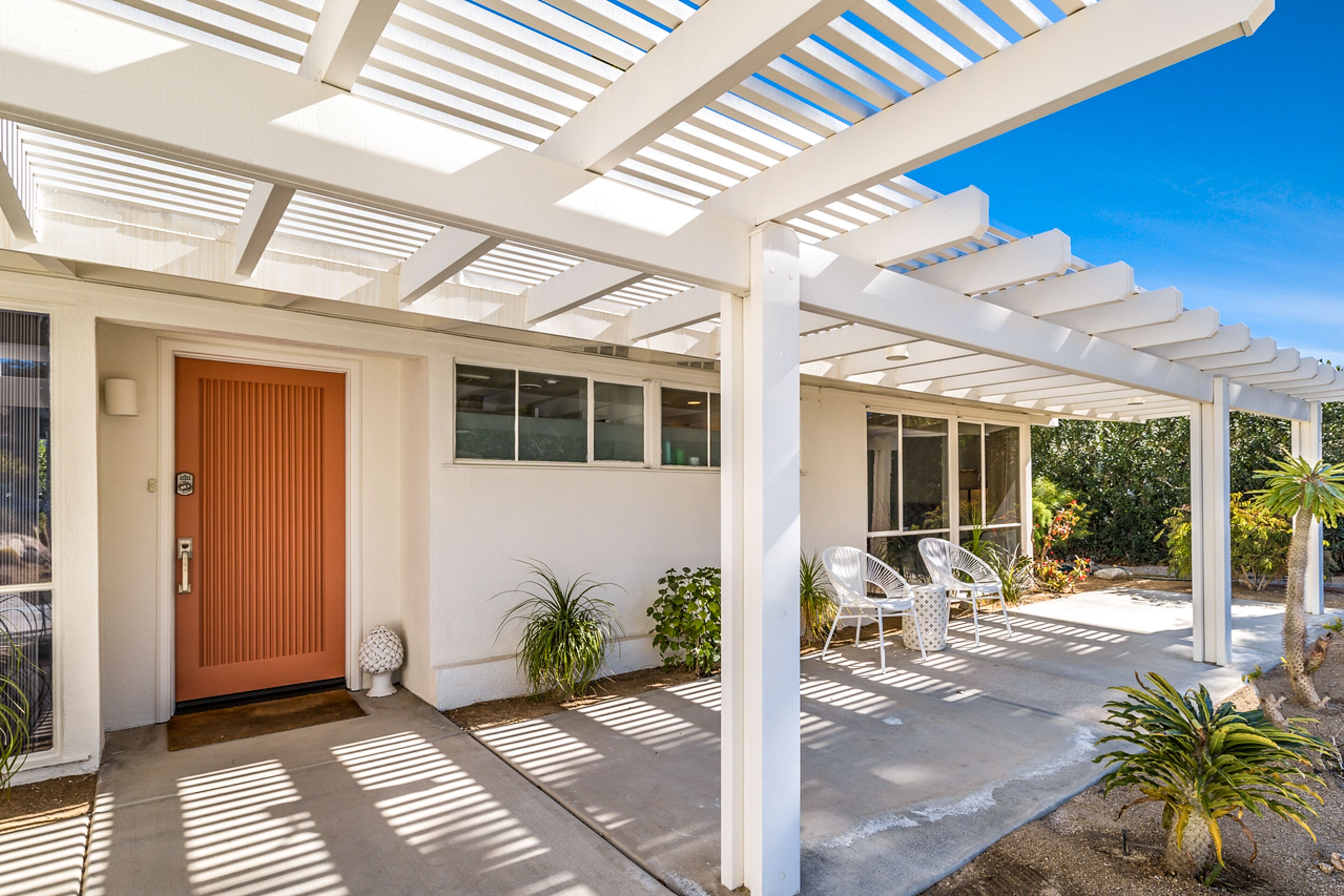 A covered patio area with a white pergola, a wooden door, and a seating area with white chairs.