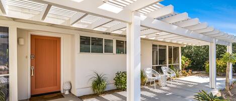 A covered patio area with a white pergola, a wooden door, and a seating area with white chairs.