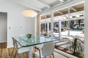 A modern dining area with a glass table and chairs, large windows, and a view of the outdoors. - Renovated, modern home with private pool, firepit, washer/dryer & AC (Rancho Mirage)