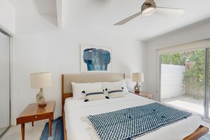 Ein modernes Schlafzimmer mit einem großen Bett, zwei Nachttischen, einem Deckenventilator und einem Fenster mit Blick auf Grünflächen.