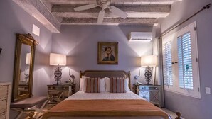 Une chambre à coucher avec un lit en bois, deux tables de chevet avec des lampes, un ventilateur de plafond, un grand miroir et une fenêtre avec des stores.