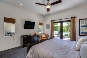 A bedroom with a large bed, a ceiling fan, a TV on the wall, a desk with a chair, and a view of the outdoors through a sliding door. - Airy, dog-friendly home with private pool, spa, & firepit - close to town (Palm Springs)