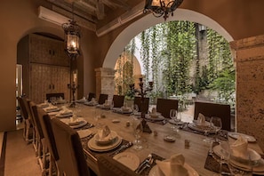 Un comedor con una larga mesa dispuesta para una comida, rodeada de sillas, y vista a la vegetación a través de un ventanal arqueado.
