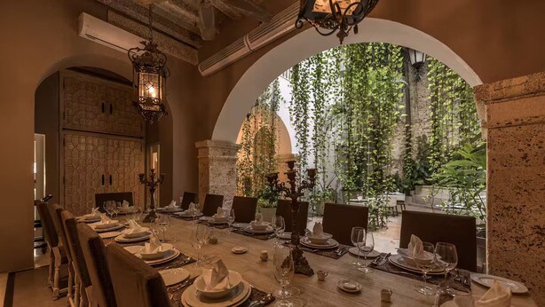 Un espace repas avec une longue table dressée pour un repas, entourée de chaises, et une vue sur la verdure à travers une fenêtre en arc de cercle.