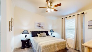Un dormitorio con cama, dos mesitas de noche con lámparas, un ventilador de techo y una ventana con cortinas.