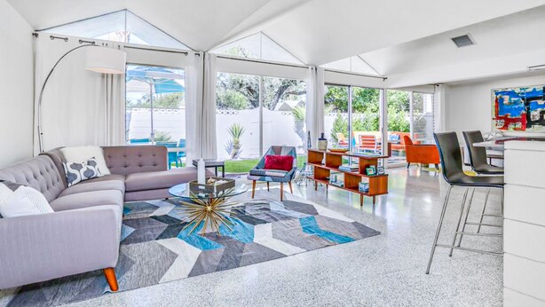 A spacious living area with a sofa, coffee table, and chairs, a kitchen area with bar stools, and large windows offering a view of the outdoors. - Charming & bright mid-century condo with shared pool - Free WiFi (Palm Springs)