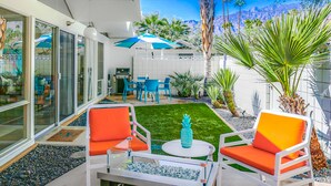 A modern outdoor patio with white furniture, a small lawn, and a dining area with blue chairs and umbrellas. - Charming & bright mid-century condo with shared pool - Free WiFi (Palm Springs)
