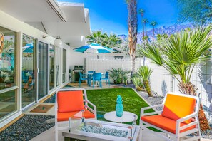 A modern outdoor patio with white furniture, a small lawn, and a dining area with blue chairs and umbrellas. - Charming & bright mid-century condo with shared pool - Free WiFi (Palm Springs)