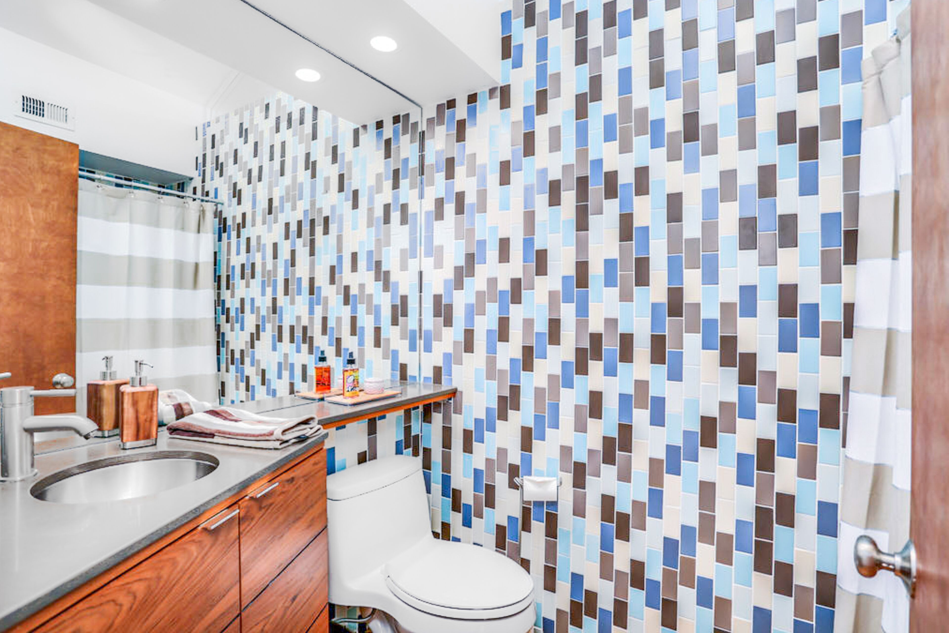 Une salle de bain moderne avec un mur décoré d’une mosaïque, un meuble-lavabo en bois, des toilettes blanches et un rideau de douche.