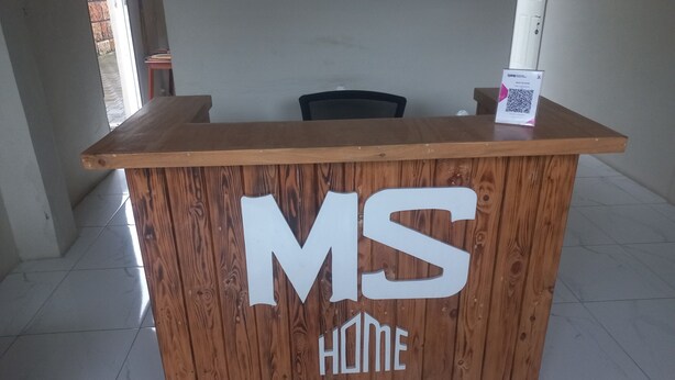 A wooden reception desk with "MS HOME" branding, a chair, and a QR code sign. - OYO 95184 Ms Home Syariah (Tegal)
