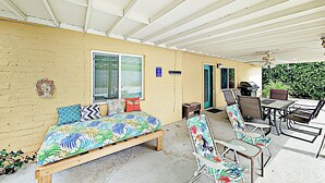 Een overdekt buitengebied met een bed, stoelen, een tafel en een barbecue.