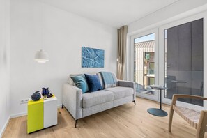 A modern living room with a grey sofa, a small white and yellow side table, a blue abstract painting, and a large window with a view of buildings. - The View B13 Fain Leewen - Balcony, elevator,shared. Sauna, parking (Dagebüll)