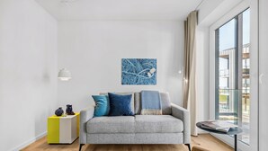 Ein modernes Wohnzimmer mit einem grauen Sofa, einem gelb-weißen Beistelltisch, einem blauen Kunstwerk und Blick durch ein großes Fenster.