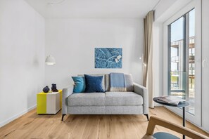 A modern living room with a grey sofa, a yellow and white side table, a blue artwork, and a view through a large window. - The View B13 Fain Leewen - Balcony, elevator,shared. Sauna, parking (Dagebüll)