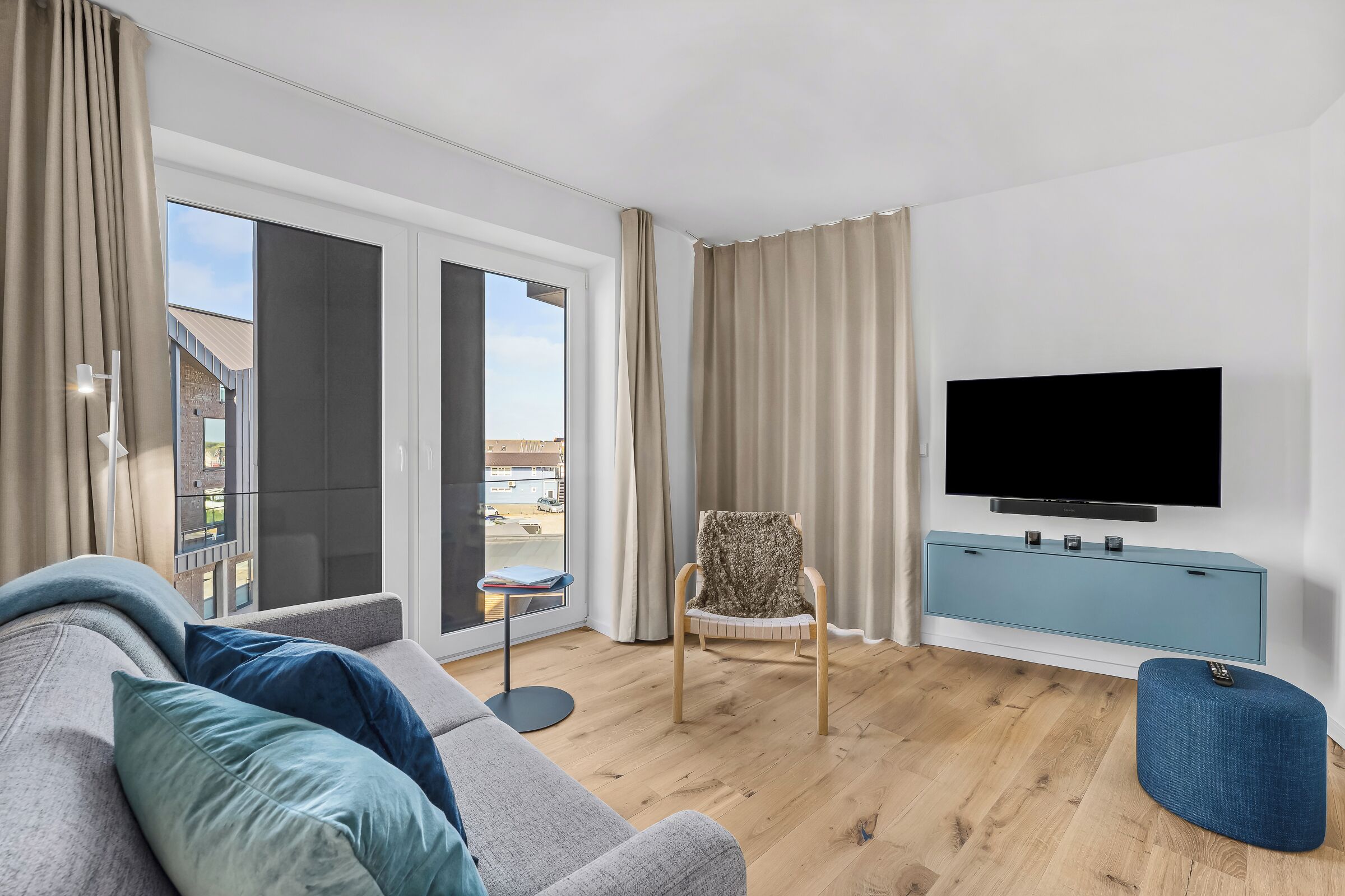 Ein modernes Wohnzimmer mit einer Couch, einem Fernseher, einem Sessel und Blick durch große Fenster.