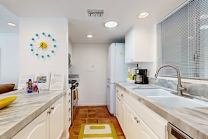 A compact kitchen with white cabinets, a marble countertop, a yellow clock, and a window with blinds. - Dog Friendly Mid-Century Villa at The Coco Cabana with Pool, Spa & Tennis Courts (Palm Springs)