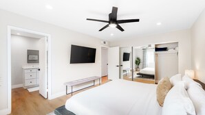 Ein modernes Schlafzimmer mit einem großen Bett, einem Deckenventilator, einem an der Wand montierten Fernseher und einem begehbaren Kleiderschrank.