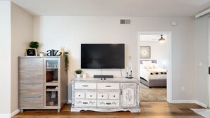 Una sala de estar con televisor, un mueble blanco con cajones, una planta pequeña y una cama al fondo.