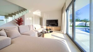 Un saló modern amb un sofà, una taula de centre i una televisió. Hi ha una escala i una vista a una piscina exterior.