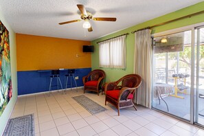 A room with a ceiling fan, a television, two wicker chairs with red cushions, a blue bar counter, and a large sliding glass door leading to an outdoor area. - Dog-friendly home with private pool, yoga studio, fitness room & great patio (Sun City)
