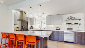 Une cuisine moderne dotée d’un grand îlot central, de tabourets de bar orange vif, d’appareils électroménagers en acier inoxydable et de luminaires suspendus.