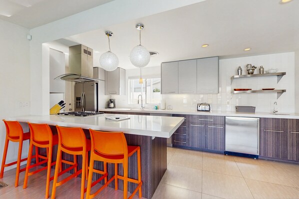 A modern kitchen with a large island, bright orange bar stools, stainless steel appliances, and pendant lighting.