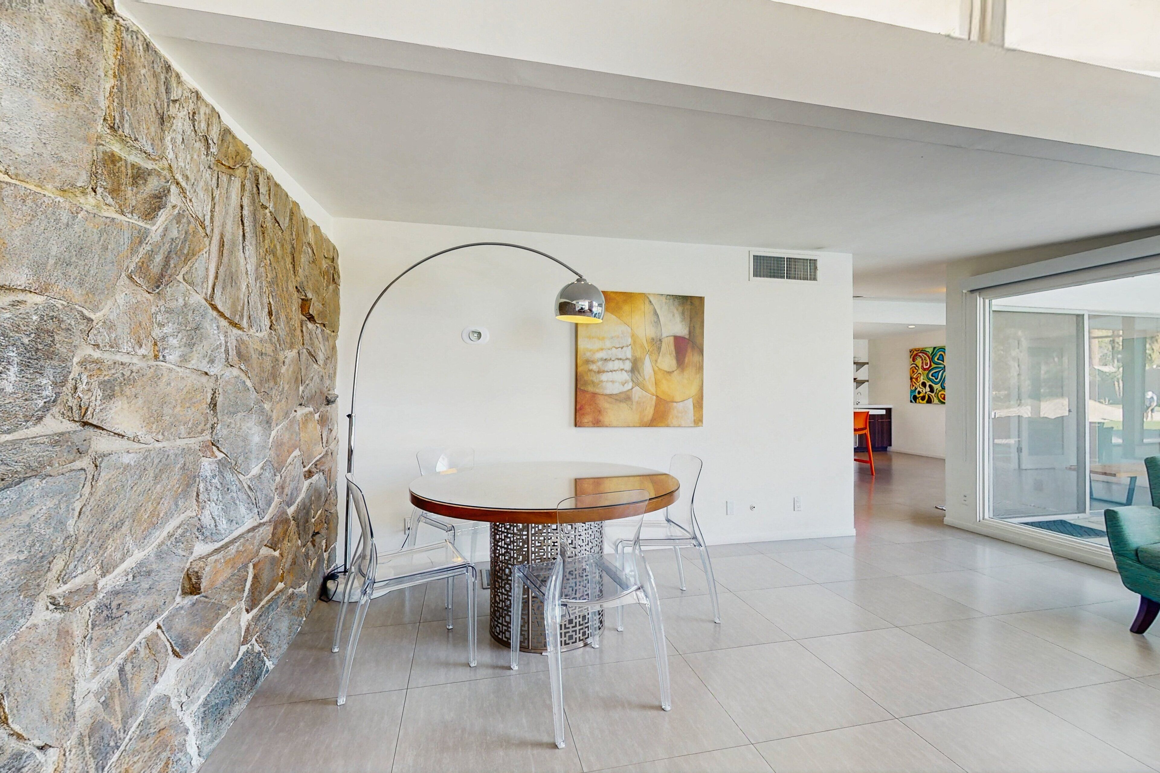 Un comedor moderno con pared de piedra, una mesa redonda, sillas traslúcidas, una lámpara colgante y un cuadro abstracto de gran tamaño.