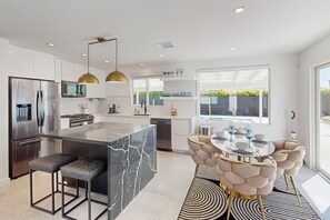 A modern kitchen with a central island, stainless steel appliances, and a dining area with a round table and tufted chairs. - Beautiful home with private pool, pool spa & open living space (Palm Springs)