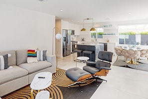 A modern living room with a sofa, coffee table, and ottoman, a dining area with a table and chairs, and a kitchen with appliances and pendant lights. - Beautiful home with private pool, pool spa & open living space (Palm Springs)