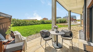 Ein möblierter Patio-Bereich mit Tisch und Stühlen, ein überdachter Sitzbereich und Blick auf ein grünes Feld sowie Gebäude im Hintergrund.