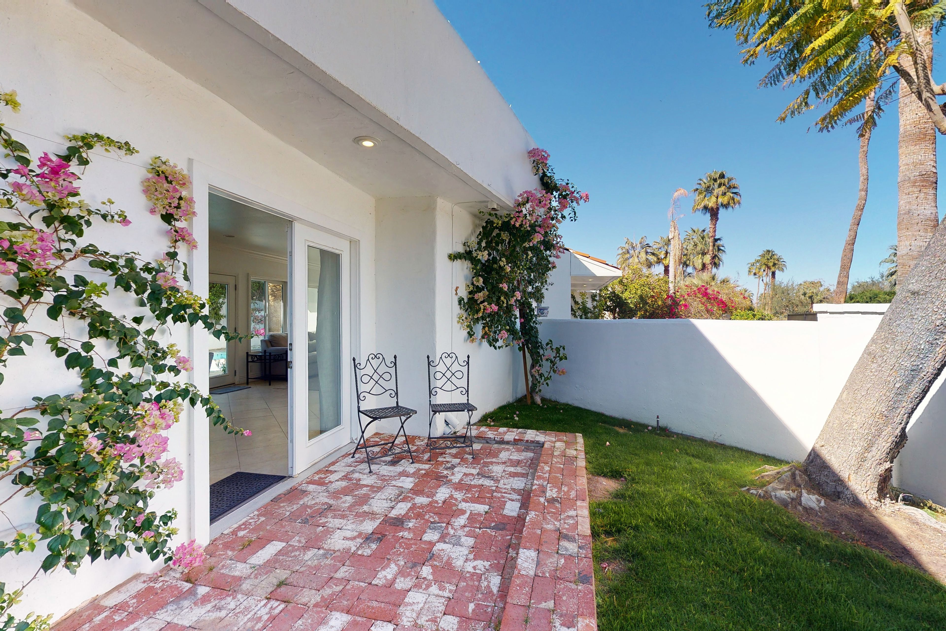 A patio with two metal chairs, a brick floor, and a white wall with climbing plants.