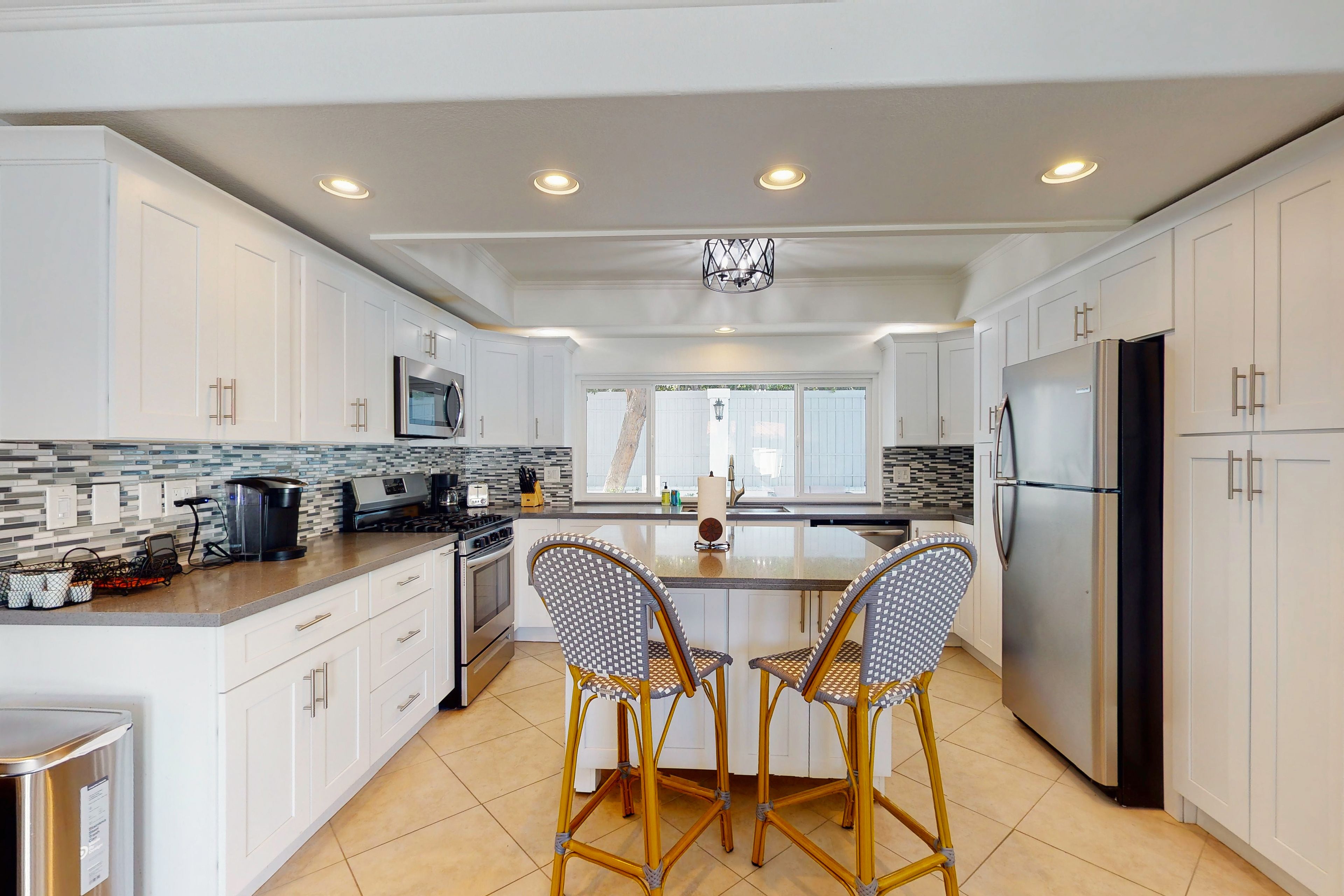 A modern kitchen with white cabinetry, a large island, two wicker chairs, and a stainless steel refrigerator.