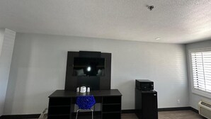 Habitació amb televisió de pantalla plana penjada a la paret, un petit altaveu negre, escriptori amb cadira i finestra amb persianes.