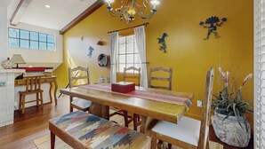 Un comedor con una mesa de madera, sillas y un banco. Hay una araña colgando sobre la mesa, y una ventana con cortinas a la izquierda.