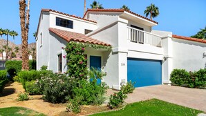 Una casa de dos pisos con puerta de garaje azul, un balcón pequeño y un jardín con diversas plantas y flores.