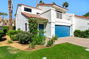 A two-story house with a blue garage door, a small balcony, and a garden with various plants and flowers. - Palm-lined mountain view condo with heated pool, tennis, grill, fireplace, & AC (La Quinta)