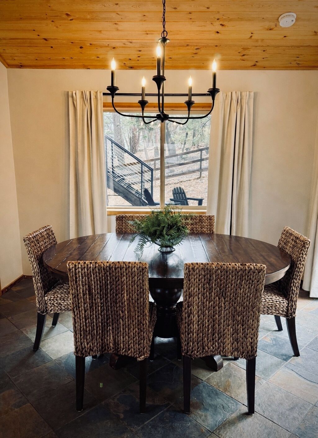 Una mesa de comedor redonda de madera con sillas trenzadas, un florero negro con follaje y un techo de madera con candelabro.