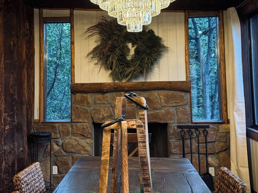 A dining area with a wooden table, chairs, a chandelier, a stone fireplace, and a wreath on the wall.