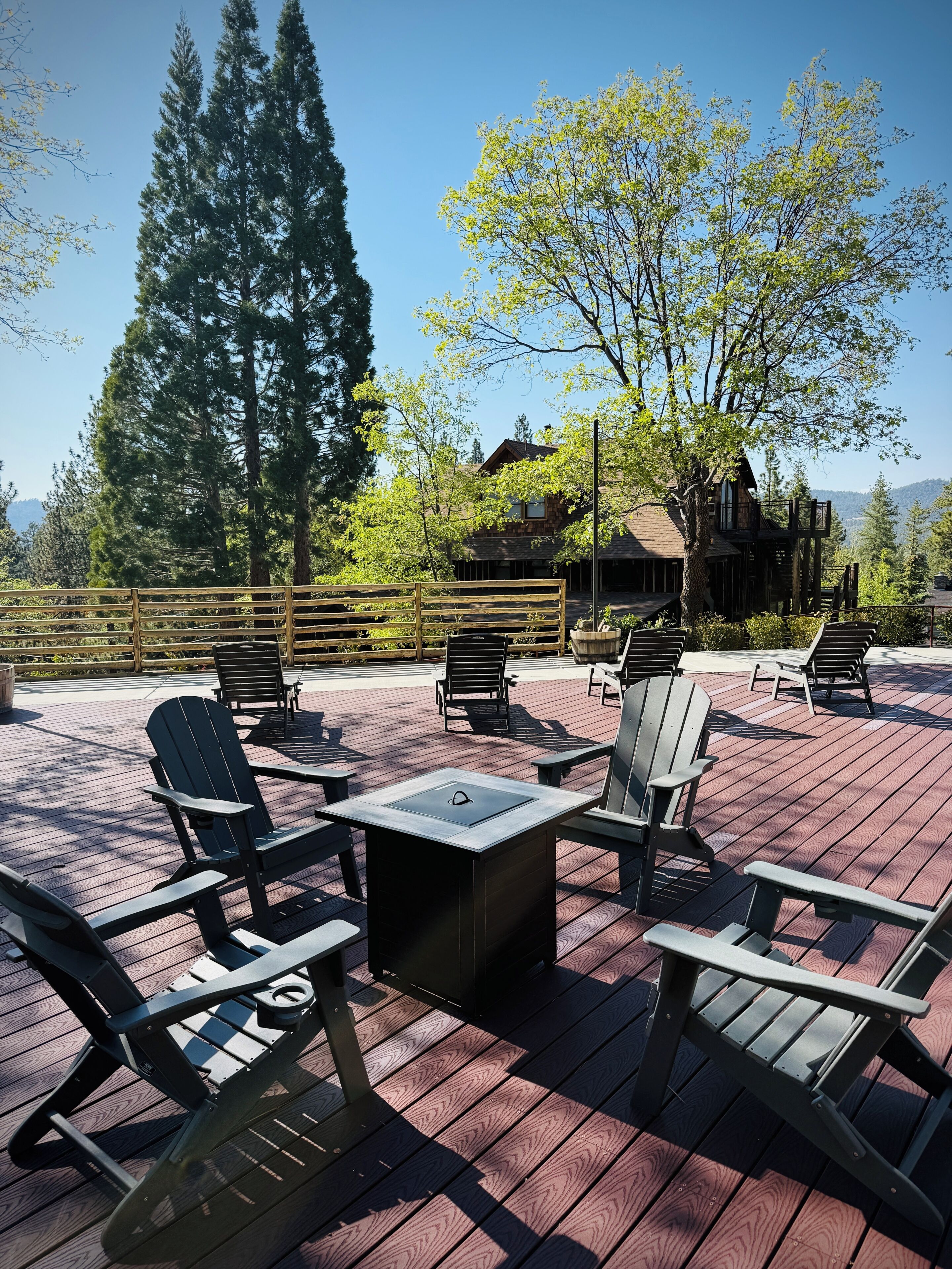 A deck with Adirondack chairs and a central table, surrounded by trees and a cabin in the background.