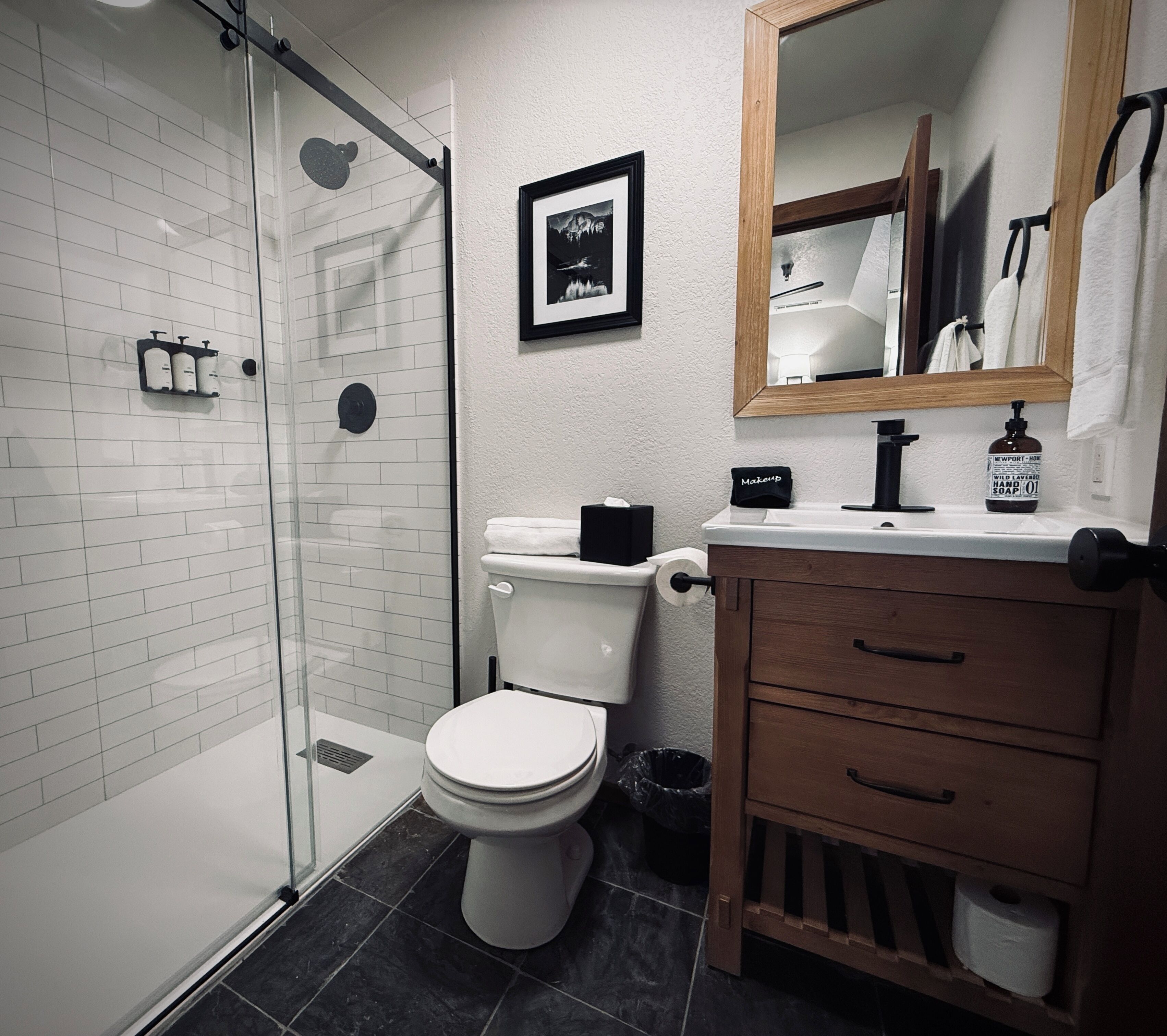 A bathroom with a glass-enclosed shower, a toilet, a vanity with a mirror, and a framed picture on the wall.