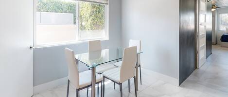 A modern dining area with a glass table and white chairs, a large window with blinds, and a stainless steel refrigerator.