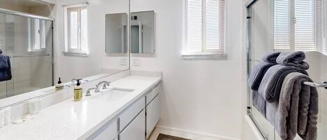 A bathroom with a white countertop, a double sink, a large mirror, and a shower area with a glass door.