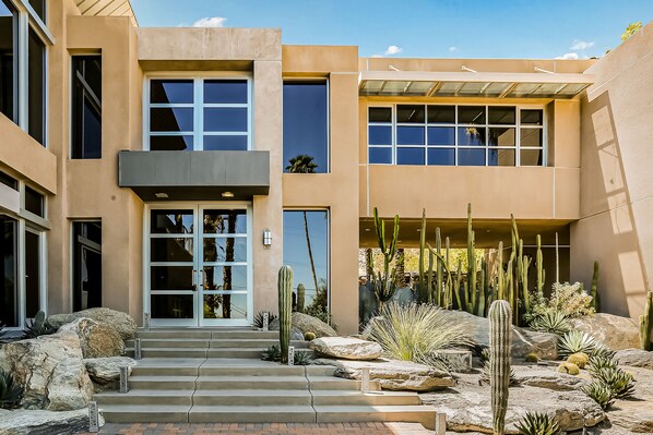 Un edificio moderno con entrada de vidrio, escalones de piedra y un jardín desértico con cactus.
